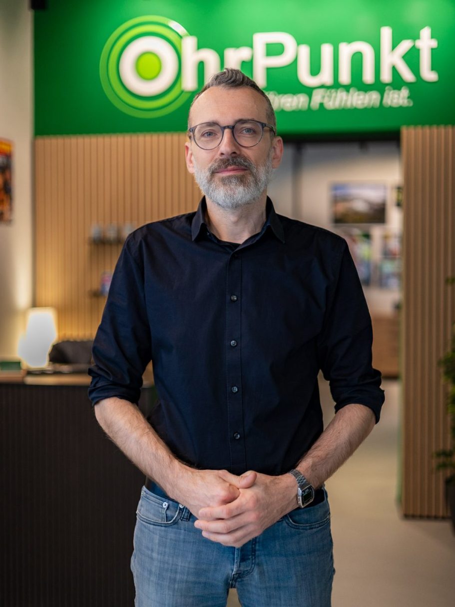Mann mit Brille und bart steht vor einem Geschäftsschild von OhrPunkt. Der Mann heißt Tobias Hirlinger, Mitgründer von OhrPunkt Hörgeräte.