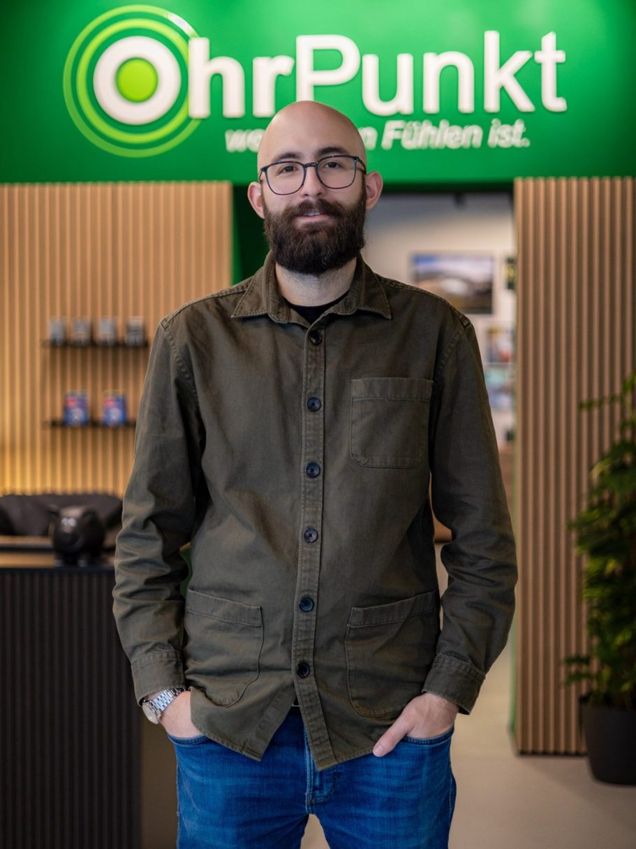 Mann mit Brille und bart steht vor einem Geschäftsschild von OhrPunkt. Der Mann heißt Andrés González, Mitgründer von OhrPunkt Hörgeräte.