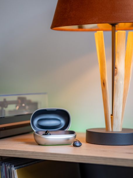 Phonak Virto R Im-Ohr-Hörsystem (IdO) neben dem kompakten Ladecase in einer modernen Lifestyle-Umgebung auf Holz.