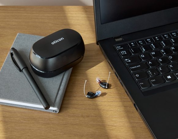 Zwei Oticon Zeal Hörgeräte neben dem kompakten Ladecase und einem Laptop in einer modernen Business-Umgebung.